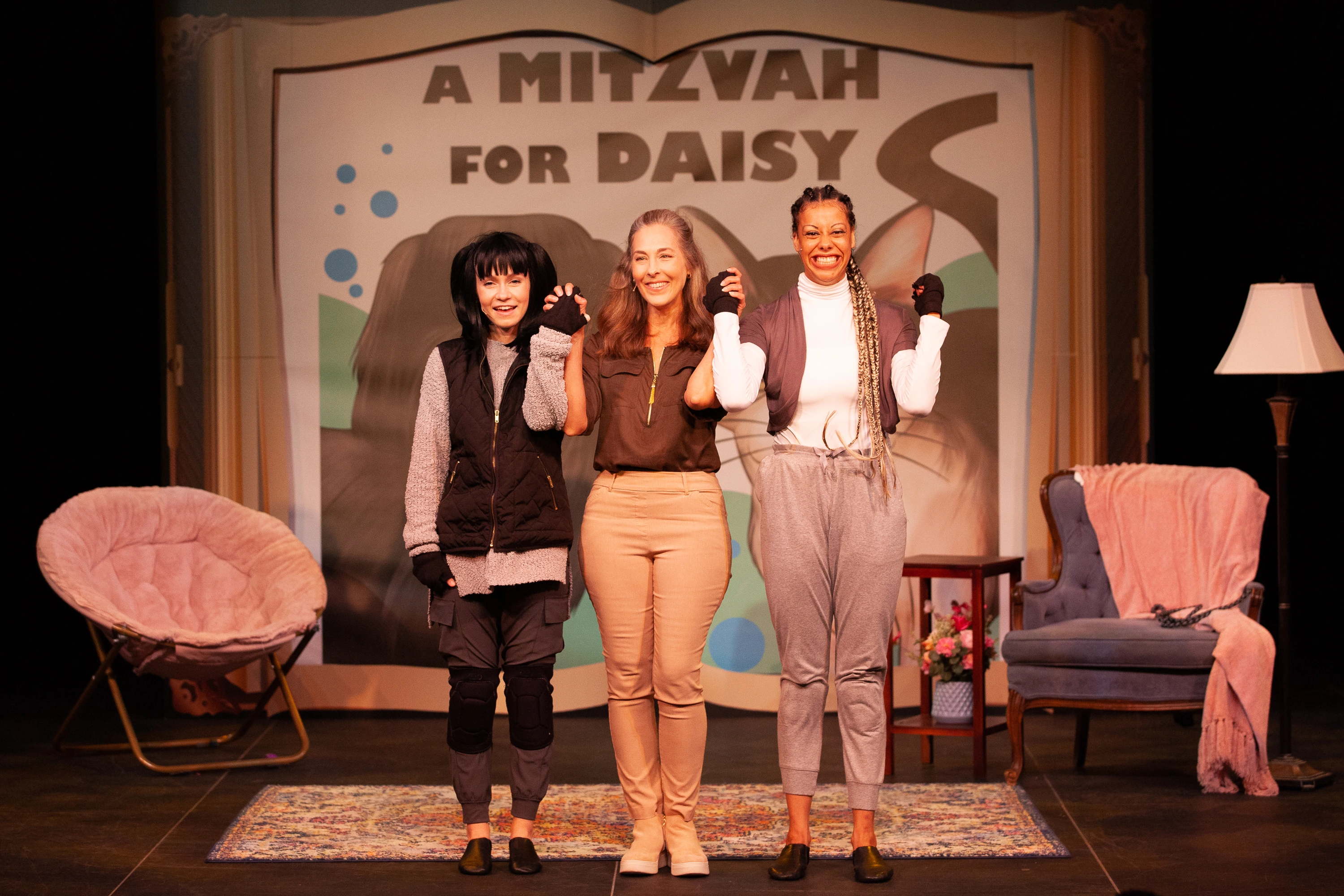 The three actors portraying Mitzvah, Mom, and Daisy stand hand in hand at center stage, smiling and taking a bow before an audience as the backdrop reads “A Mitzvah for Daisy.”