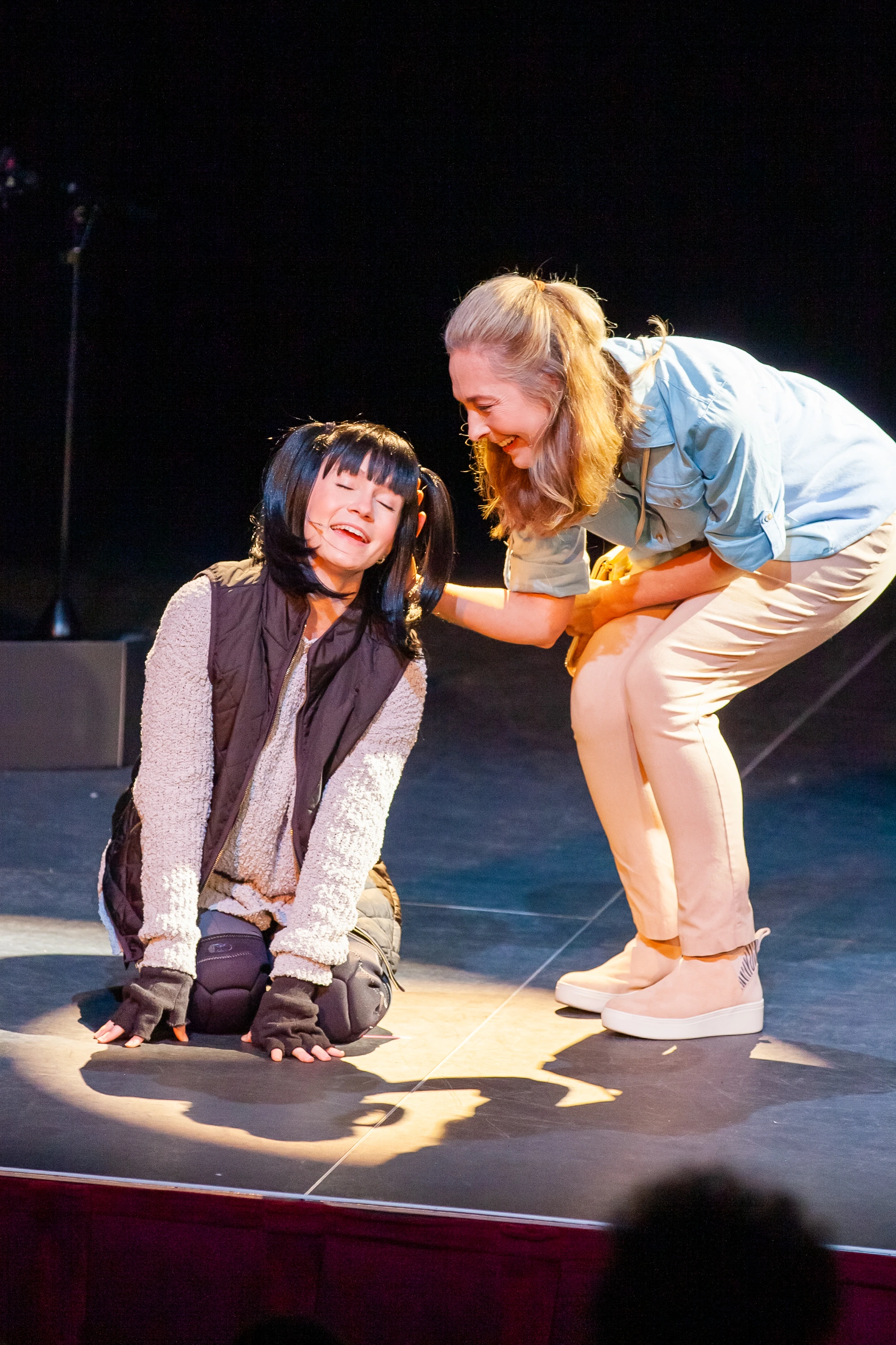 On stage, Mom gently pets Mitzvah, who kneels happily with eyes closed, smiling in contentment as they share a heartfelt moment under the spotlight.