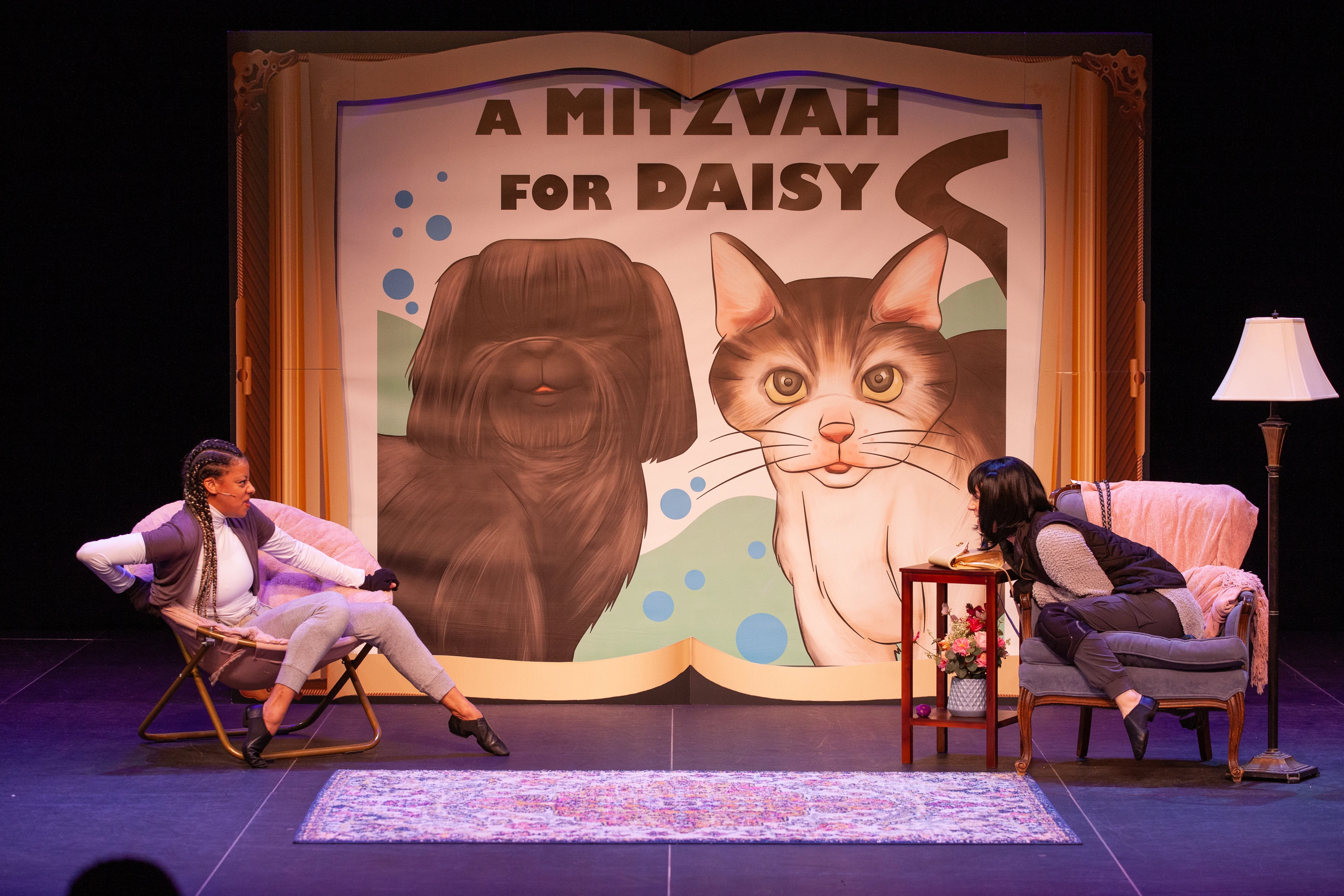 Two actors perform on stage in front of a large illustrated backdrop reading “A Mitzvah for Daisy,” showing a dog and a cat. One actor lounges in a chair while the other leans forward from an armchair, portraying Daisy and Mitzvah adjusting to each other.