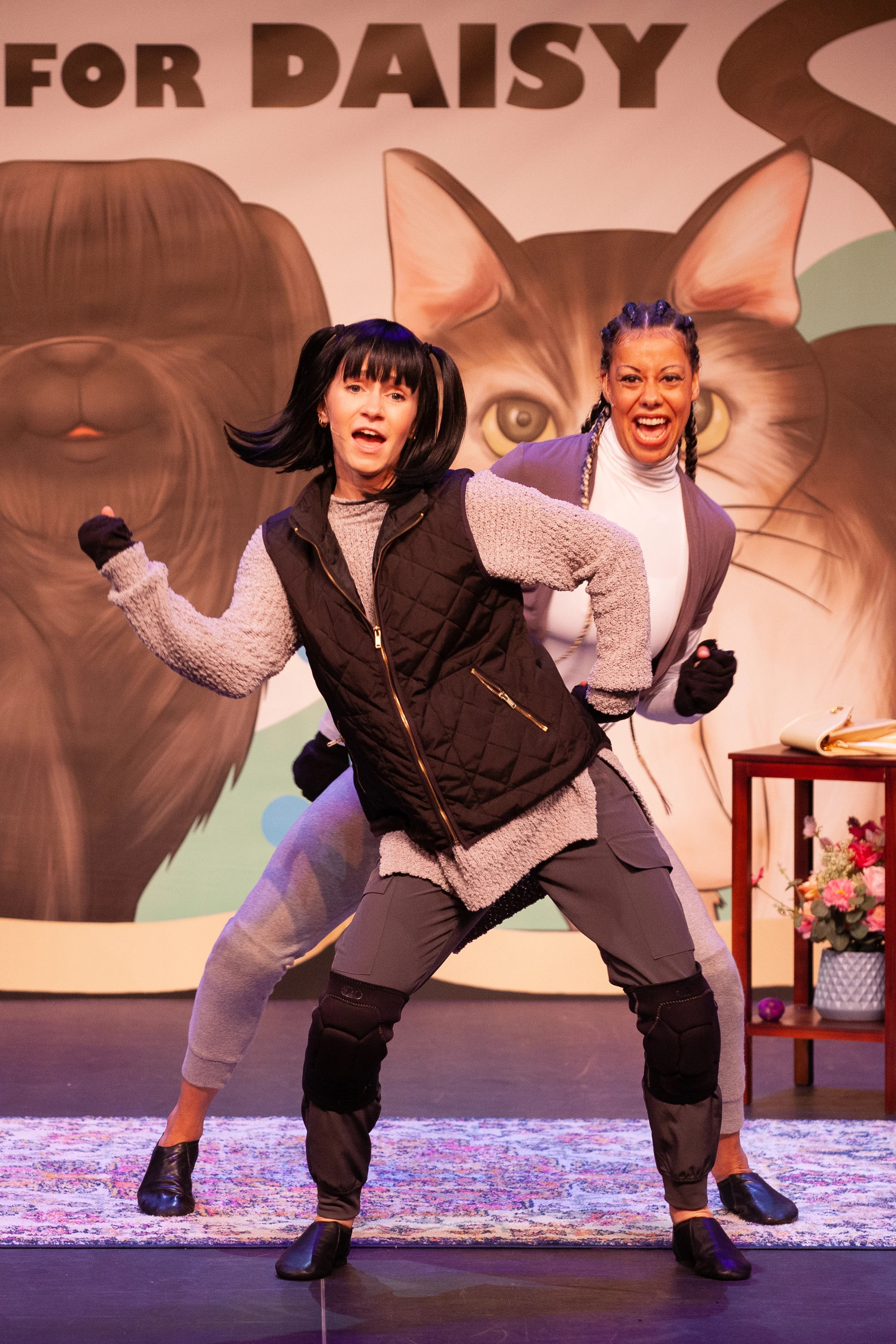 Two actors portraying Mitzvah and Daisy stand on stage mid-dance in front of a colorful backdrop reading “A Mitzvah for Daisy,” featuring large painted portraits of a dog and a cat. Both performers smile joyfully, celebrating the friendship between the two characters.