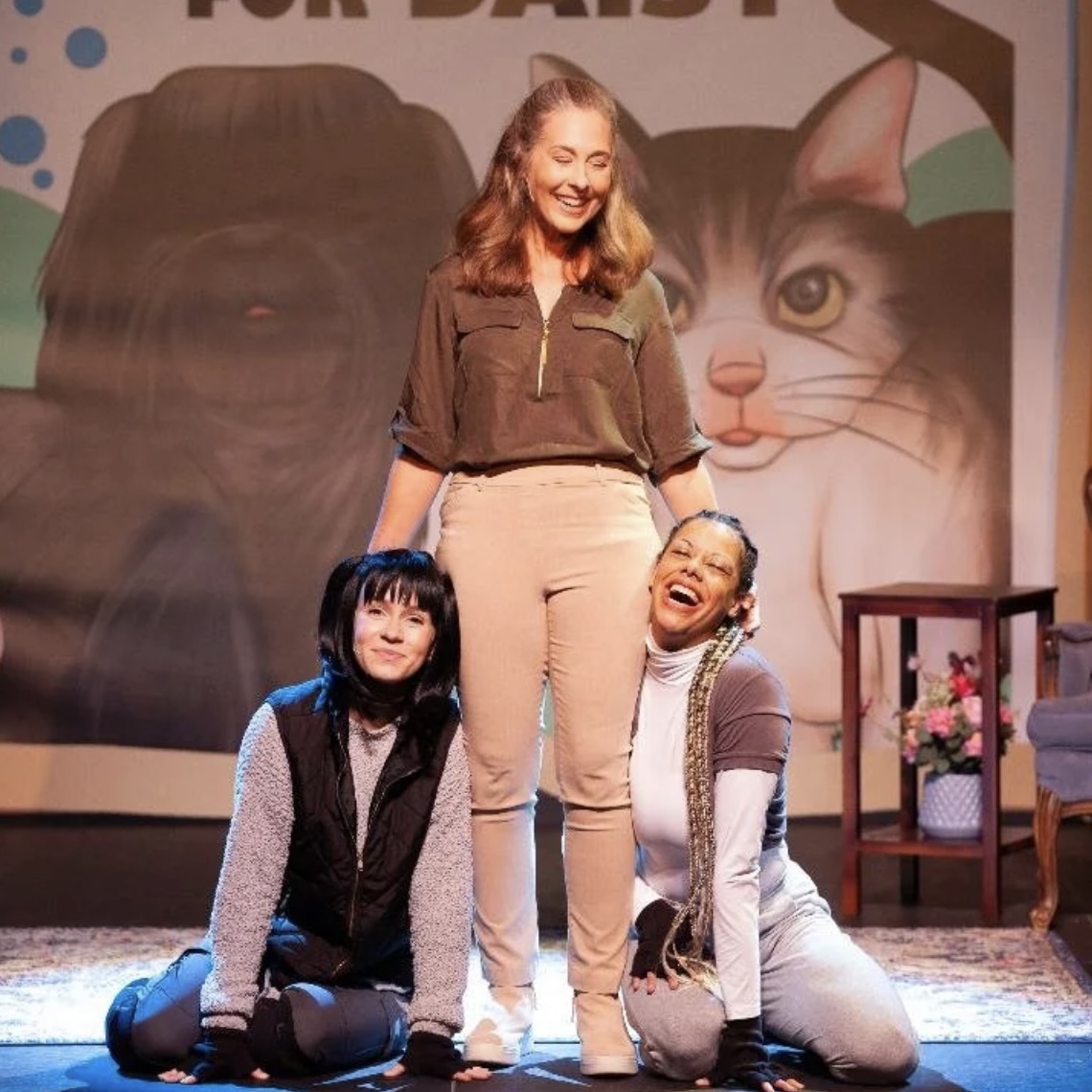 “Three actors on stage smiling after a performance of A Mitzvah for Daisy, with one standing at center and two seated at her feet in costume, in front of a large illustrated backdrop of a dog and cat.