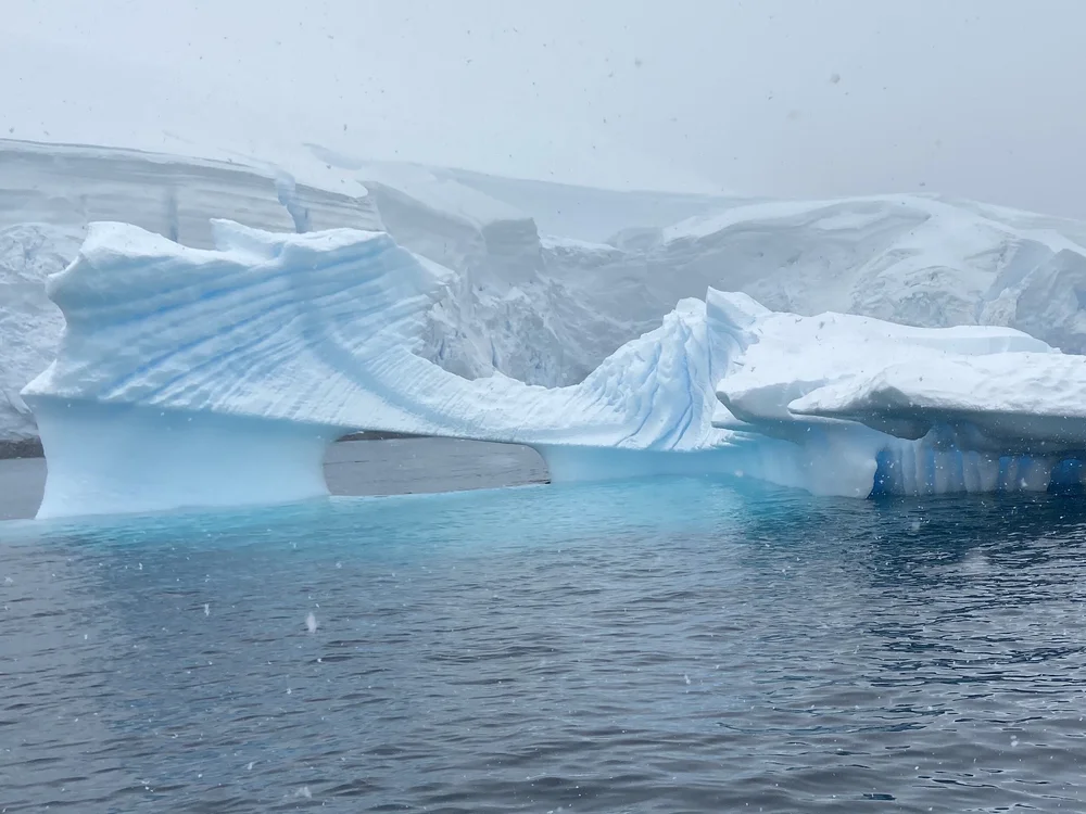 Sculpted iceberg with an arch and bright blue ice floating in Antarctic waters on a snowy day
