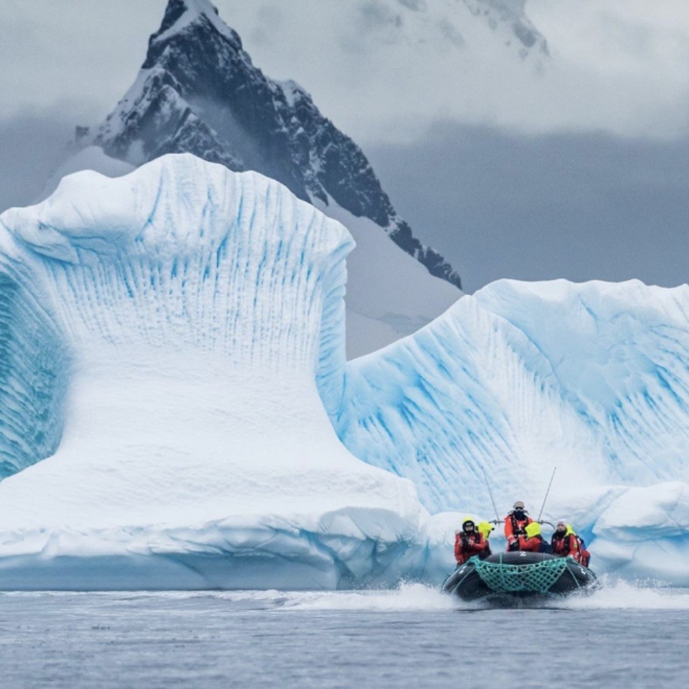  A group of people in red jackets ride a small inflatable boat past massive blue icebergs, with a dark jagged mountain peak rising behind them in Antarctica.