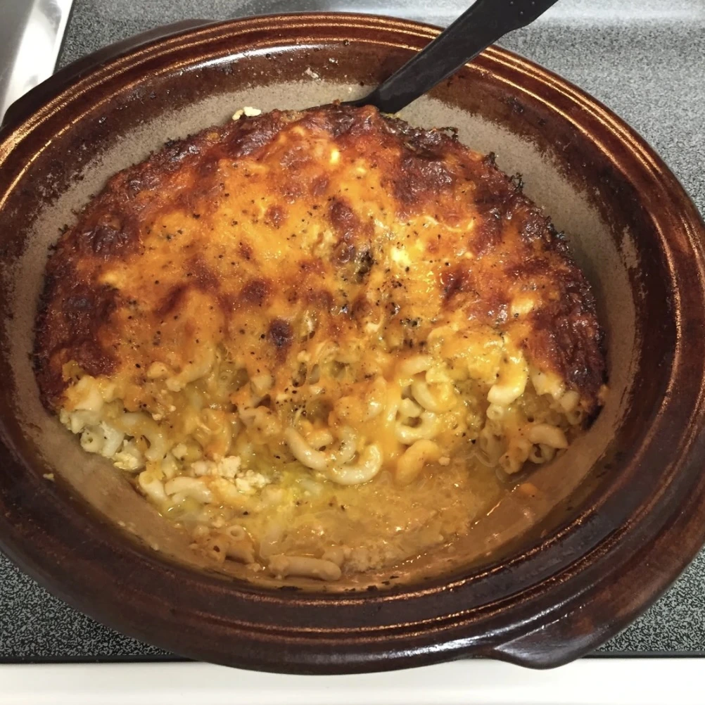 A dish of baked macaroni and cheese with a golden, crispy cheese crust on top, partially scooped out to reveal creamy noodles underneath, sitting on a stovetop.