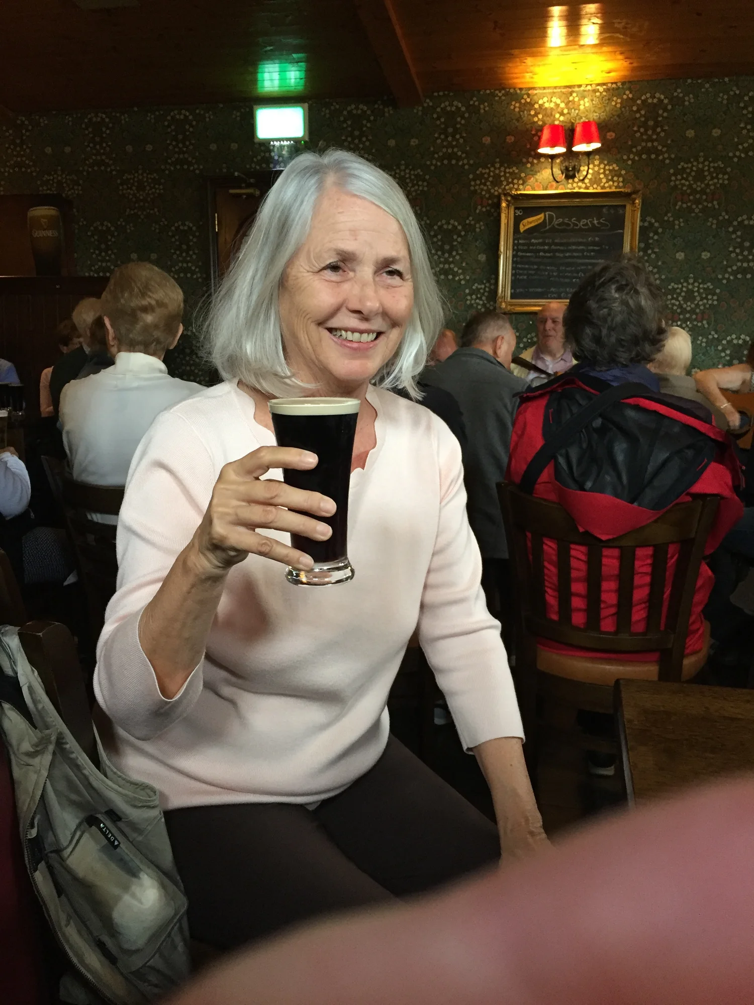 Deborah Lee Prescott smiling and holding a pint of dark beer inside a lively pub during her travels in Ireland, surrounded by other patrons.
