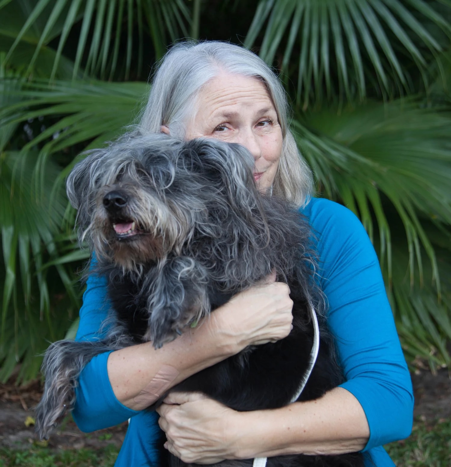Deborah Lee Prescott smiles while hugging Mitzvah, the fluffy black dog who inspired her children's book. Lush green palm fronds fill the background.
