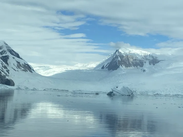 Snow-covered Antarctic mountains with a glacier flowing into calm reflective waters under a partly cloudy sky