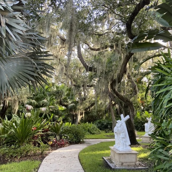 A peaceful garden path winds through lush greenery and moss-draped trees, lined with white statues depicting the Stations of the Cross.