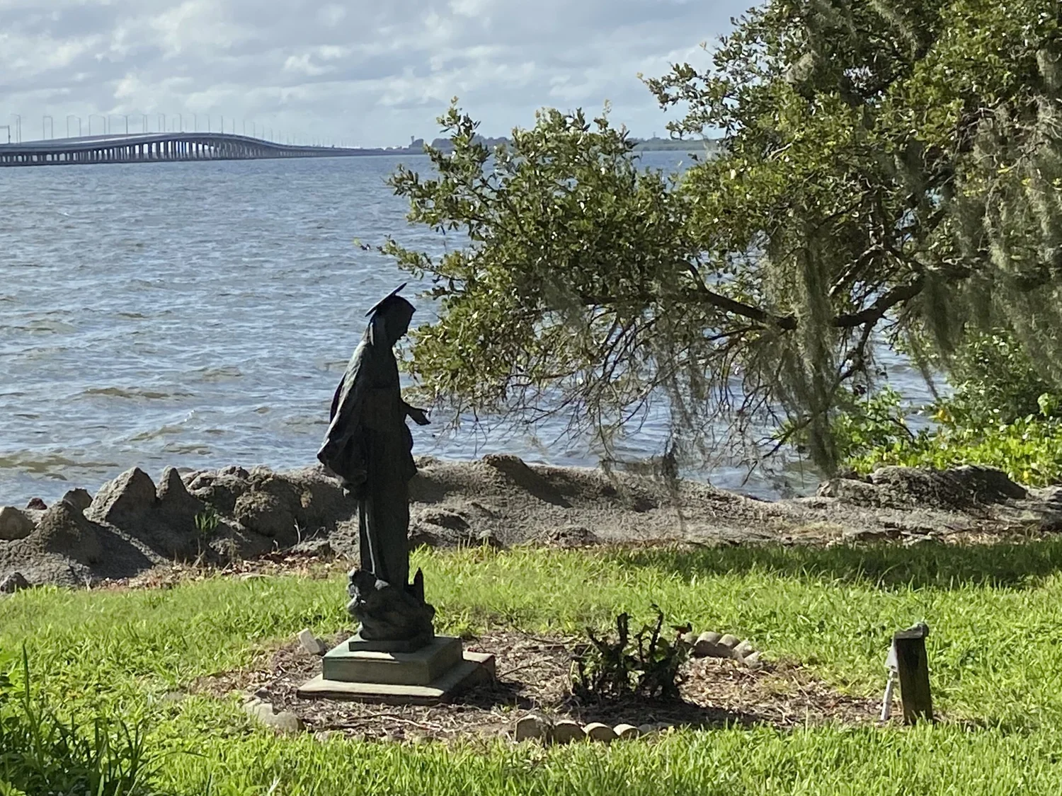 Statue of a robed figure near the shoreline, with a tree partially overhanging and a bridge visible in the distance across the water.