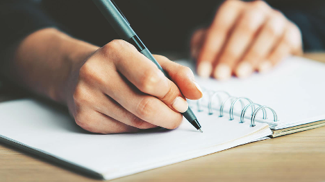 Close-up of a hand writing with a pen in a spiral notebook, symbolizing journaling or memoir writing.